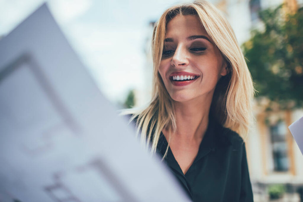 Happy female in casual clothing analyzing information from papers sitting in sidewalk cafe, carefree hipster girl with blonde hair doing journalistic research holding documents and smiling - Фото, изображение