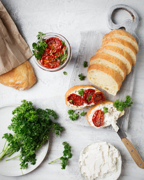 Italienische Küche. Bruschetta mit getrockneten Tomaten und Quark. Italienisches Essen auf weißem Hintergrund. Brot in Scheiben auf einem Brett. Toasts werden vorbereitet. Getrocknete Tomaten und frische Kräuter. Baguette - Foto, Bild