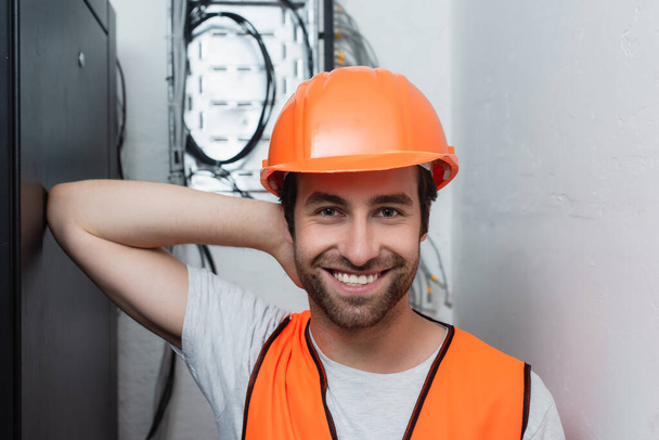 Young serviceman in hard hat standing near blurred switchboard  - Foto, imagen