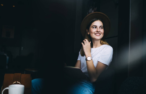 Jeune femme souriante positive dans un chapeau élégant et des vêtements à la mode assis au café avec ordinateur portable tout en regardant loin et toucher les cheveux - Photo, image