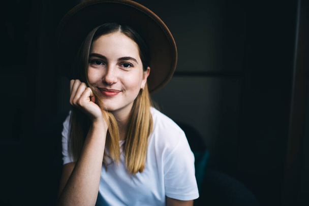 Positiivinen hymyilevä nuori nainen tyylikäs hattu ja rento vaatteita käsin leuka istuu nojatuolissa ja katselee kameraa - Valokuva, kuva