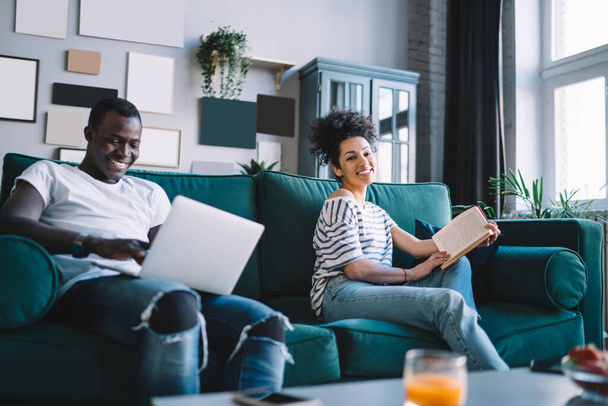 Curly ethnic female in casual clothes smiling while sitting with interesting book on cozy sofa near African American male using laptop at home - Zdjęcie, obraz