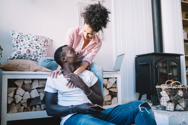 Ethnic young woman sitting in room with fireplace and embracing from back black man sitting on floor with smartphone while telling story - Foto, Bild
