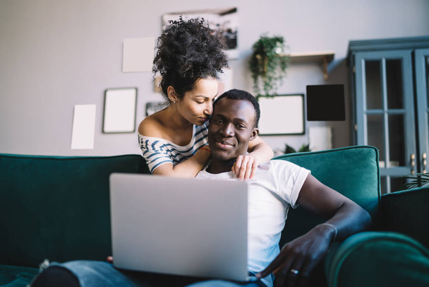 Young African American female with closed eyes embracing and touching black male while spending time at home and browsing laptop together - Photo, image