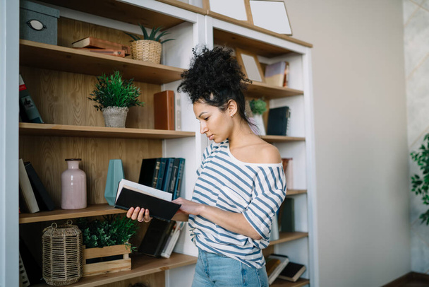 Charming smart ethnic woman holding open book in black cover nearby bookshelves and looking through pages in light living room - Foto, afbeelding