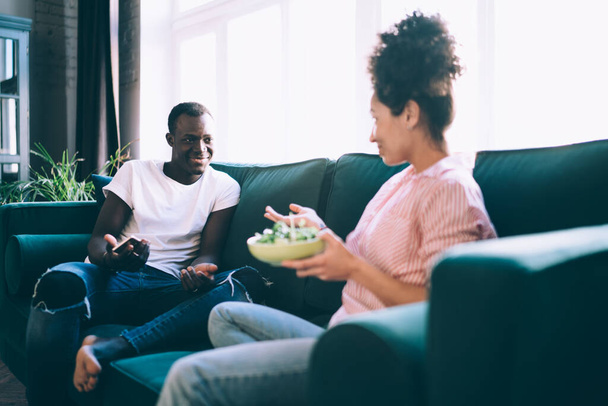 Diverse young happy girlfriend and boyfriend in casual clothes talking and having fun while sitting on comfortable sofa with meal  - Фото, изображение