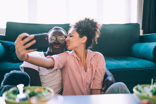 Happy multiracial adult people in casual clothes taking selfie with smartphone while sitting on floor and kissing against green sofa in light modern apartment - Foto, afbeelding