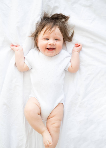 funny baby girl in white bodysuit smiling and lying on a white bedding at home. top view. - Foto, imagen