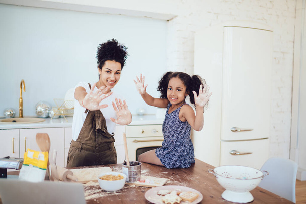 Smiling ethnic pretty mother and little cute daughter showing hands stained with flour at camera while girl helping mom baking in modern kitchen at home - Fotoğraf, Görsel