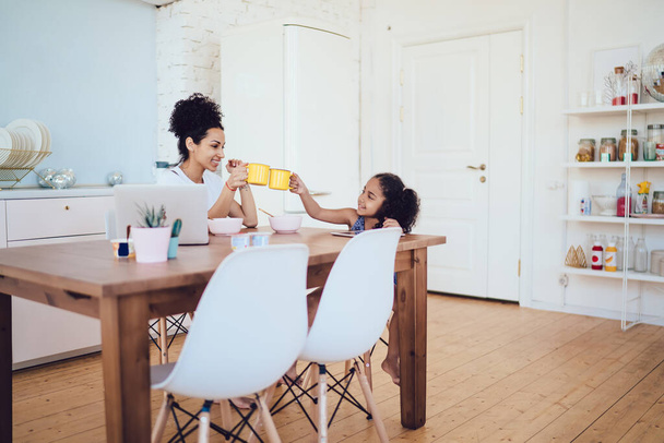 若いですアフリカ系アメリカ人巻き女性と小さな娘toastingとともに朝食マグカップ座っていますテーブルで居心地の良いキッチンお互いを見ます  - 写真・画像