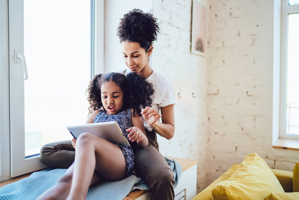 Brunette African American girl with disgusted face lying and using tablet with mother on windowsill near bright window in cozy room - Valokuva, kuva