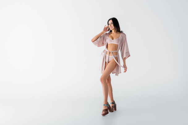 full length of cheerful young woman posing in underwear and silk robe on white - Foto, afbeelding