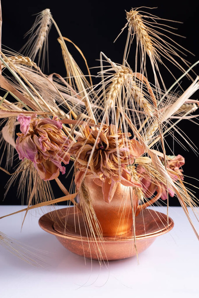 a bouquet of wheat ears with dry flowers in a copper dish on a white table and black background. - Фото, зображення