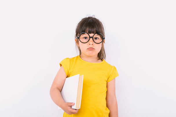 Niña con gesto serio usando camiseta amarilla y gafas negras redondas sosteniendo un papel blanco en una mano sobre fondo blanco - Foto, imagen