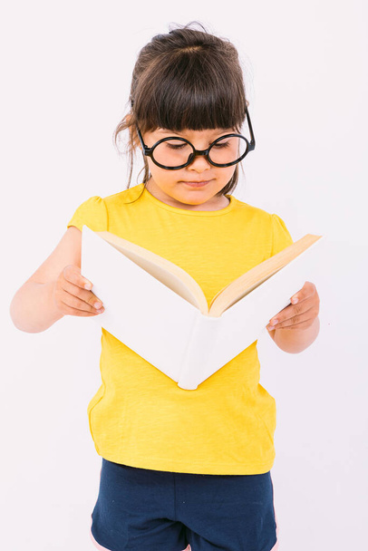 Mosolygó kislány, sárga pólóban és kerek fekete szemüvegben, nyitott könyvet tartva a kezében és olvasva, fehér alapon - Fotó, kép