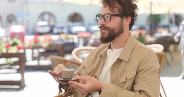 joven casual con anteojos, sentado en una terraza, bebiendo café y sintiéndose satisfecho con él - Metraje, vídeo