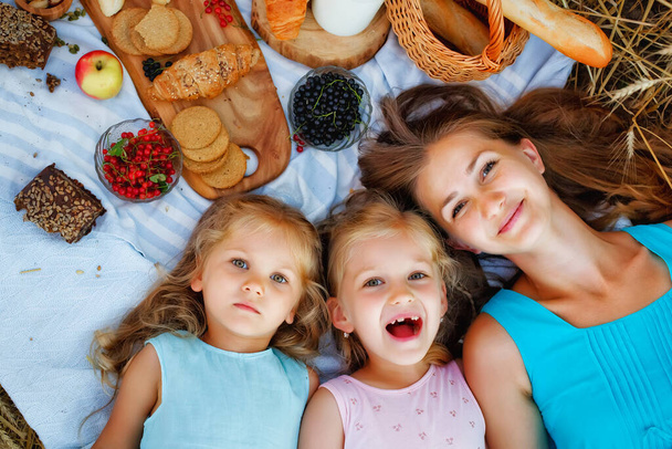 Πορτρέτο μιας ευτυχισμένης οικογένειας σε πικνίκ. Μια μητέρα και δύο κόρες είναι ξαπλωμένες σε μια κουβέρτα δίπλα σε μια ξύλινη σανίδα με φαγητό. Άνω όψη. - Φωτογραφία, εικόνα