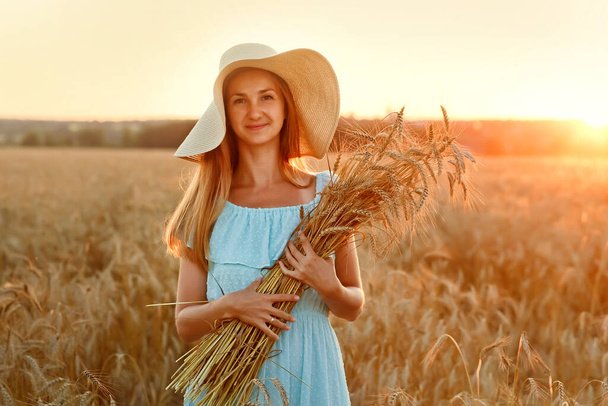 Une jeune femme tient des épis de maïs dorés dans un champ de blé. Une femme avec un chapeau et une robe au coucher du soleil. L'idée d'une récolte riche - Photo, image