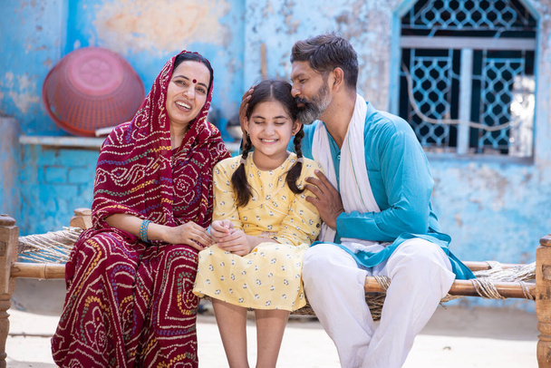 Boldog vidéki indián család mosolyog, miközben ül a hagyományos ágyban a falu otthonában, apa csókolja aranyos kislánya a fejét, és anya száriban nézi kamera szülő támogatja a szeretet gyermek. - Fotó, kép