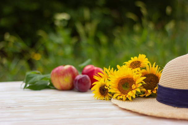 a bouquet of sunflowers and a straw hat on a background of fresh fruits on a wooden table in the garden - Fotó, kép