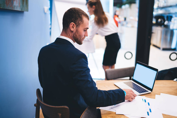 Вид збоку серйозного бізнесмена в чорному формальному костюмі з використанням ноутбука за столом, слухаючи презентацію ділового партнера в сучасному робочому просторі
 - Фото, зображення
