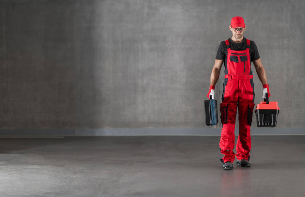 Kaukasischer Bauarbeiter in seinen Vierzigern in roter Uniform spaziert mit Werkzeugkoffern in der Hand durch die Innenräume roher Betonbauten. - Foto, Bild