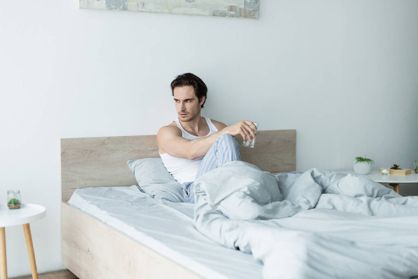 sfrustrowany mężczyzna siedzi w łóżku ze szklanką wody i odwraca wzrok - Zdjęcie, obraz