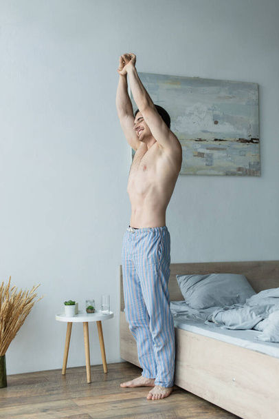 glimlachende man in gestreepte pyjama broek stretching met gesloten ogen in slaapkamer - Foto, afbeelding
