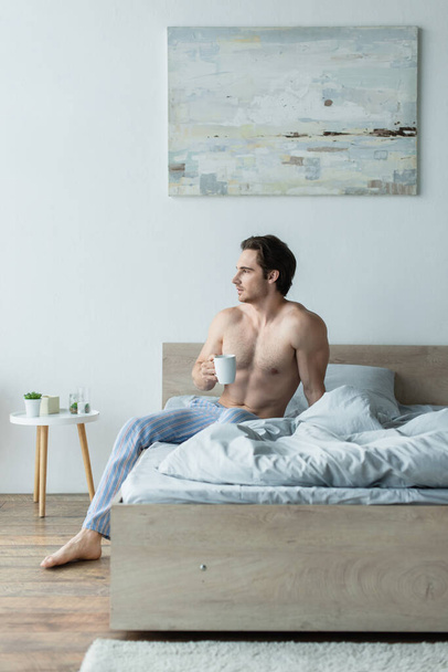 shirtloze man die wegkijkt terwijl hij op bed zit met een kop koffie - Foto, afbeelding