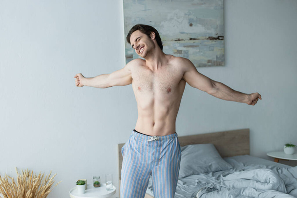 shirtless man in pajama pants smiling and stretching in bedroom - Foto, Imagem