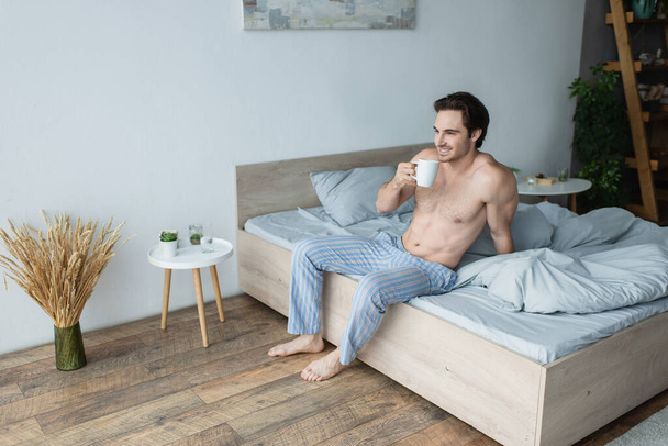 χωρίς πουκάμισο άντρας με ριγέ παντελόνι πιτζάμας χαμογελώντας πίνοντας καφέ στην κρεβατοκάμαρα - Φωτογραφία, εικόνα