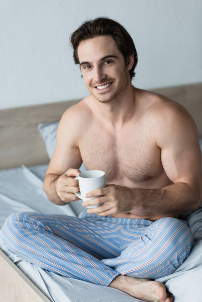 shirtloze man lachend naar de camera terwijl hij in bed zit met een kop koffie - Foto, afbeelding