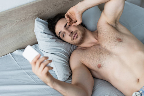 young man looking at blurred smartphone while rubbing eye in bed - Foto, imagen
