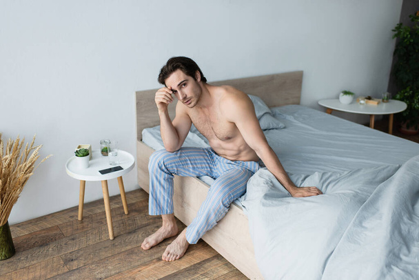 shirtloze man in blauwe pyjamabroek die zich onwel voelt terwijl hij 's morgens op bed zit - Foto, afbeelding