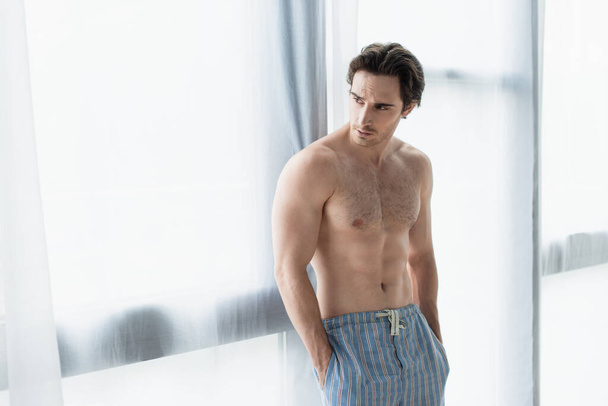 shirtless man standing near window with hands in pockets of pajama pants - Zdjęcie, obraz