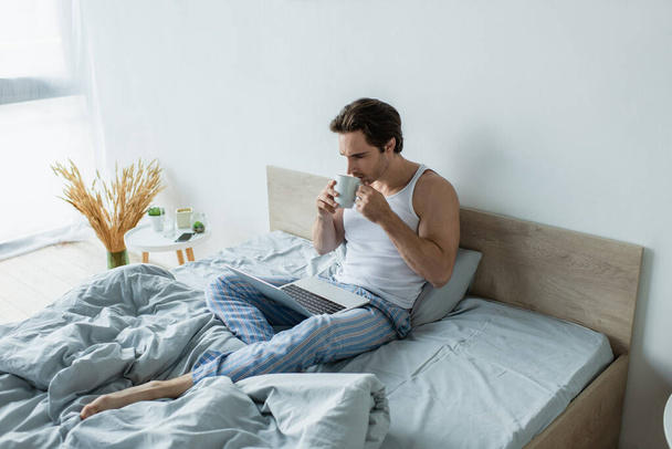young man looking at laptop while drinking coffee in bed - Fotografie, Obrázek