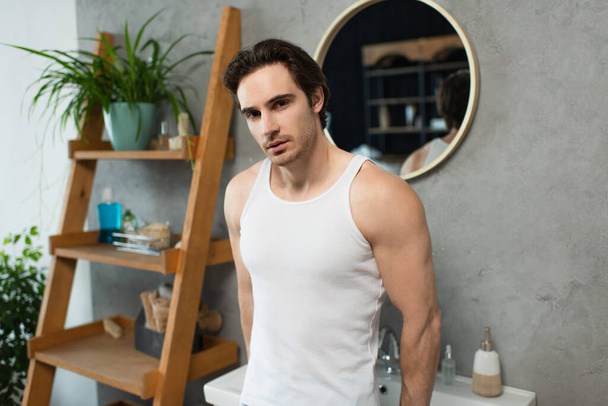 brunette man in white tank top standing near blurred rack in bathroom - Photo, Image