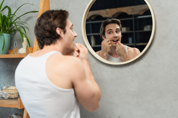 brunette man in white tank top cleaning teeth in bathroom near mirror - Fotó, kép