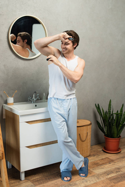smiling man in white tank top and blue pajama pants applying deodorant on armpit - 写真・画像