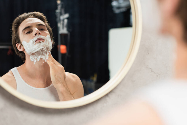brunette man aanbrengen scheerschuim op gezicht terwijl het kijken in spiegel - Foto, afbeelding