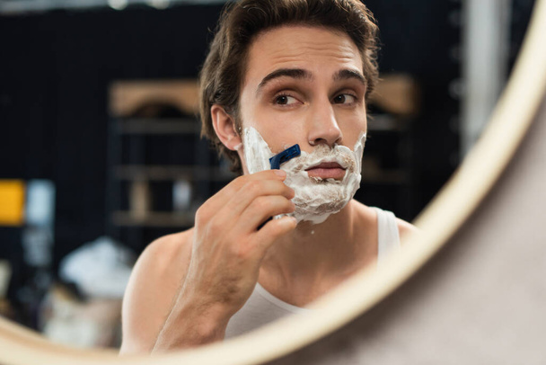 молодой брюнетка мужчина смотрит в зеркало во время бритья утром - Фото, изображение