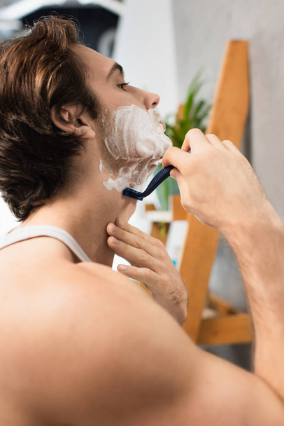 Брюнетка мужчина бритья с безопасной бритвой в ванной комнате - Фото, изображение