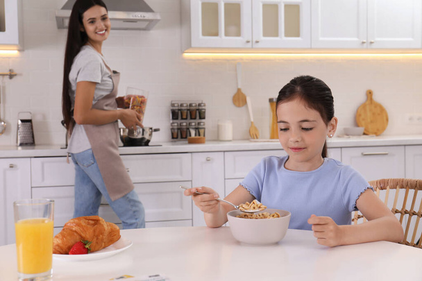 Little girl having breakfast while her mother cooking food in kitchen. Single parenting - Fotografie, Obrázek