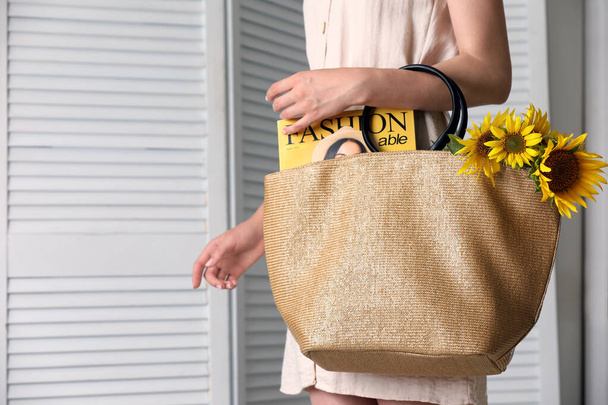 Woman holding beach bag with magazine and beautiful bouquet of sunflowers indoors, closeup - Фото, зображення