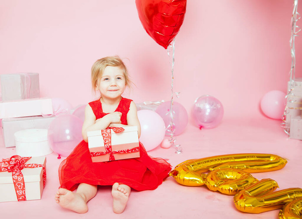 Porträt eines dreijährigen Mädchens in rotem Kleid auf rosa Hintergrund mit Geschenken und Luftballons. - Foto, Bild