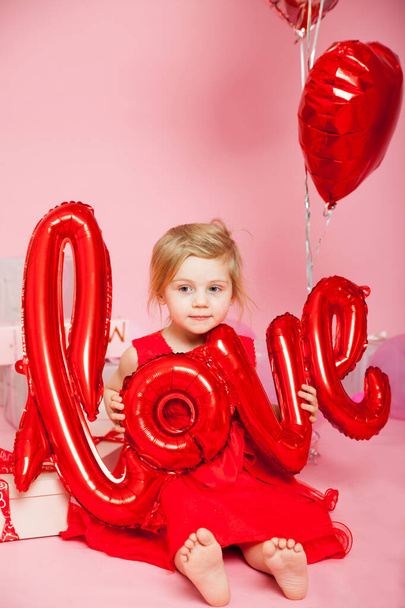 Πορτρέτο του τρίχρονου κοριτσιού με κόκκινο φόρεμα σε ροζ φόντο με μπαλόνια σε σχήμα λέξης αγάπη.  - Φωτογραφία, εικόνα