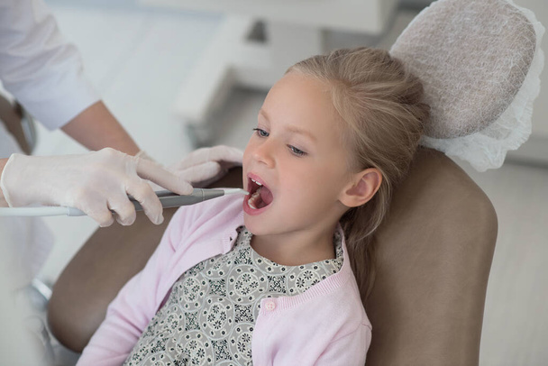 Una doctora rubia sosteniendo un taladro dental mientras trabaja con su pequeña paciente - Foto, imagen