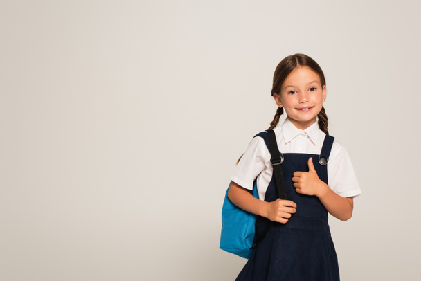 happy schoolgirl with blue backpack showing thumb up isolated on grey - 写真・画像
