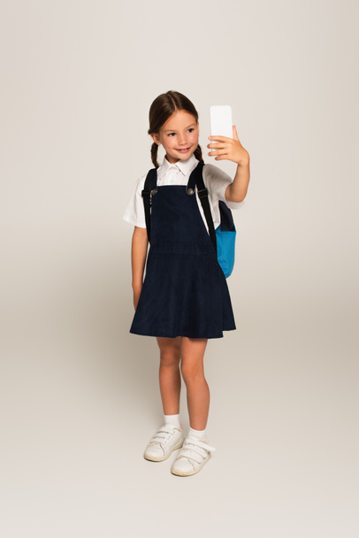 vista completa de la chica en uniforme escolar tomando selfie en el teléfono móvil en gris - Foto, imagen