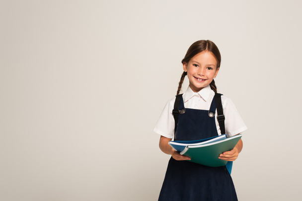 positive schoolchild smiling at camera while holding notebooks isolated on grey - Foto, afbeelding